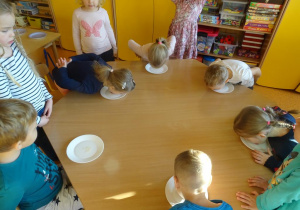 Dzieci siedzą przy stoliku jedzą miód z talerzyków bez użycia rąk. Reszta dzieci obserwuje.